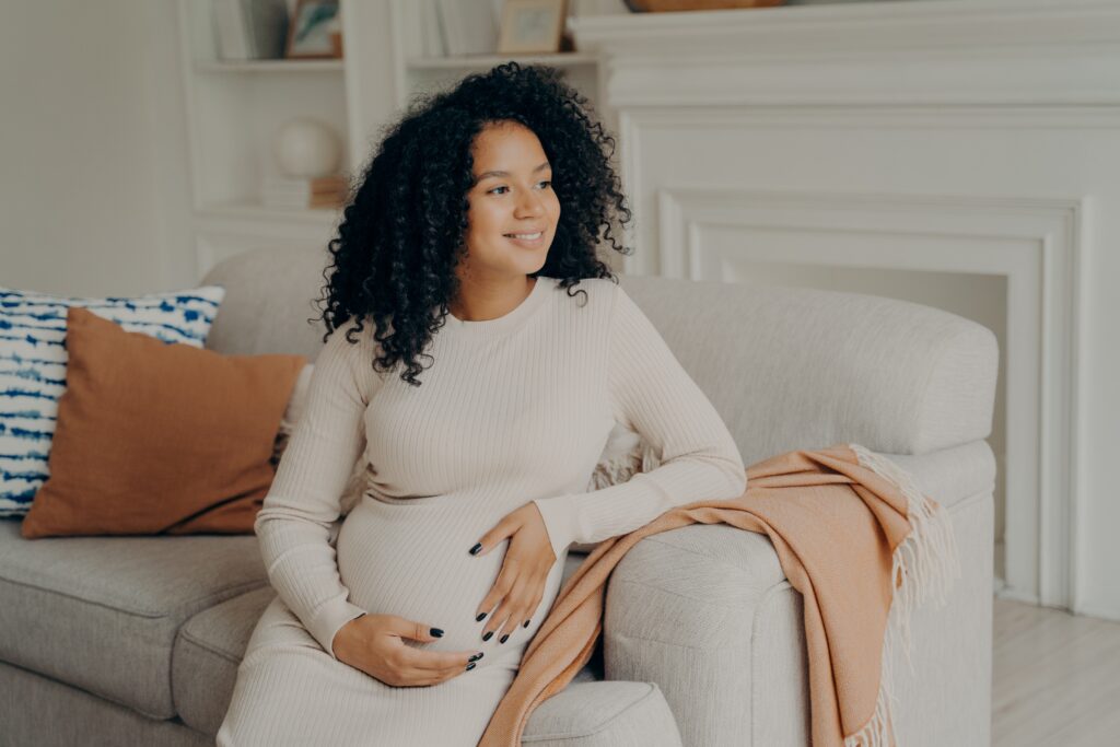 A beautiful young pregnant woman with mixed race heritage is hugging her tummy while sitting on a light-colored couch in front of a light-colored background.