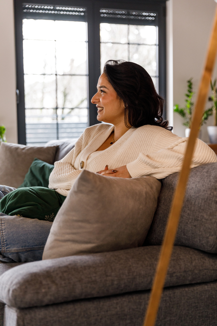 Portrait of happy young woman sitting on the cozy sofa in her living room, simply relaxing, looking away, smiling and contemplating.